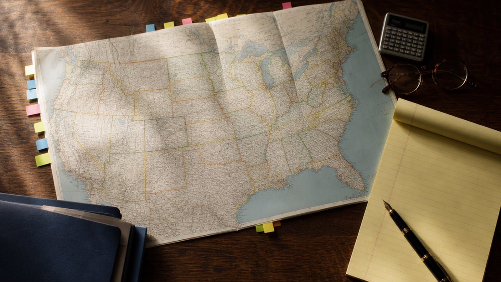 A printed United States map on a desk with sticky note tabs, a legal pad and reading glasses